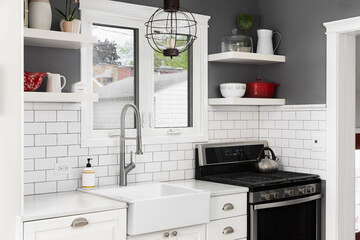 A kitchen faucet detail with white cabinets, farmhouse sink in front of a window, floating shelves on a dark grey wall, and a white subway tile backsplash.