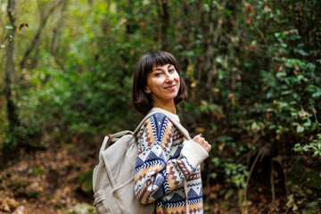 girl in a stylish sweater and with a backpack enjoys a walk in the forest. trekking in the forest. a girl with a backpack walks in the forest. trekking and hiking.