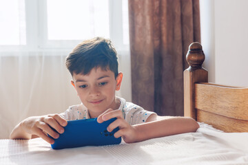 A young boy, aged 6-8, plays a game on a smartphone in a cozy room. The photo highlights themes of...
