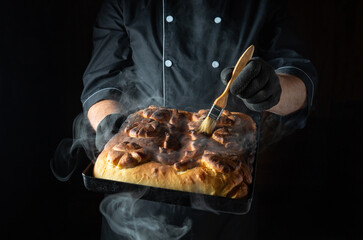 In a dimly lit kitchen, a chef skillfully applies a glaze to a steaming tray of warm pastry,...