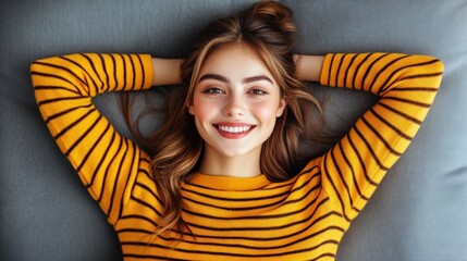 Fototapeta premium Young woman relaxing with a smile on her face, wearing a striped yellow sweater, lying on a soft gray couch