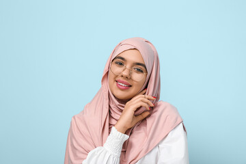 Beautiful young Muslim woman in hijab and eyeglasses on blue background