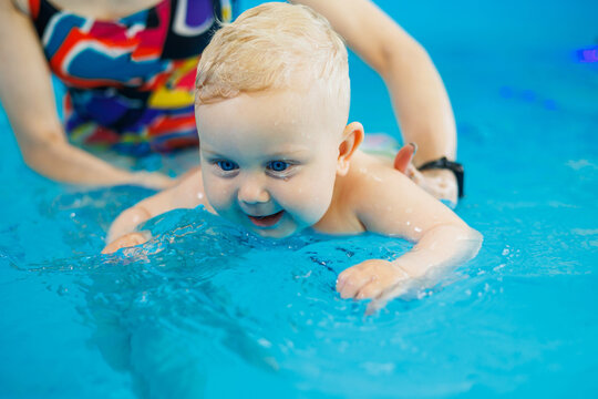 A baby learns to swim with a coach in the pool. Swimming in an early age pool. Happy child with female trainer in indoor swimming pool playing and having fun.