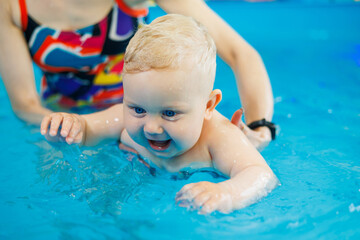 A baby learns to swim with a coach in the pool. Swimming in an early age pool. Happy child with female trainer in indoor swimming pool playing and having fun.