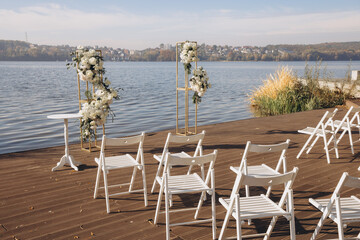 Wedding ceremony setup with white chairs and flower decorations on a wooden pier overlooking a lake