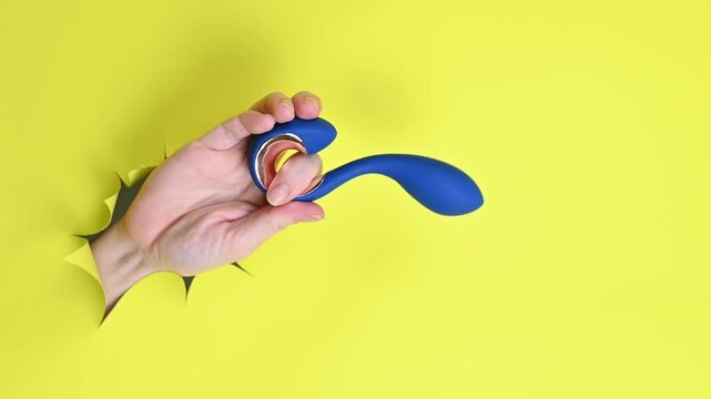 Female hand holding blue vibrator sticking out of yellow paper background. Copy space.