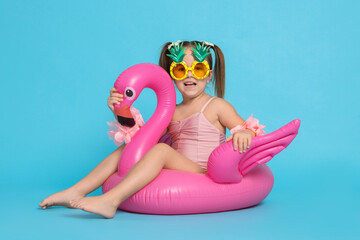 Cute little girl in swimsuit with funny sunglasses and inflatable ring on light blue background