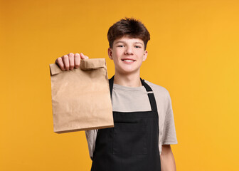 Teenage boy with paper bag working as cashier on orange background