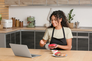 African-American woman with laptop cooking tasty eclairs in kitchen