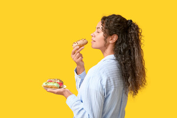African-American woman eating tasty eclairs on yellow background