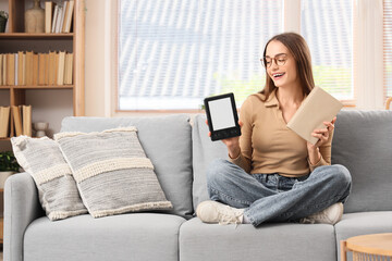 Young girl choosing between e-reader and book while sitting on sofa at home
