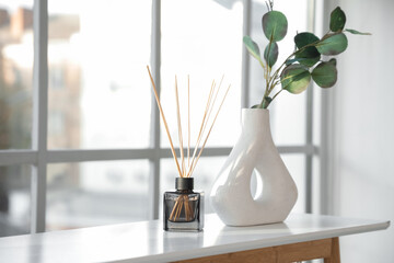 Reed diffuser and vase with eucalyptus branches on table near window in living room, closeup