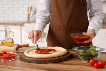 Woman spreading tomato paste on dough for pizza near table in kitchen