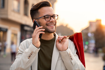 Cheerful Caucasian professional in beige blazer talks on smartphone while carrying red shopping bag...