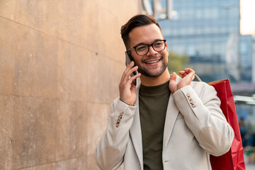 Asian professional in light blazer smiles while talking on smartphone and carrying red shopping bag...