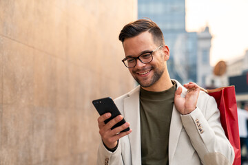 Caucasian male professional in white blazer checks smartphone while carrying shopping bag on city...