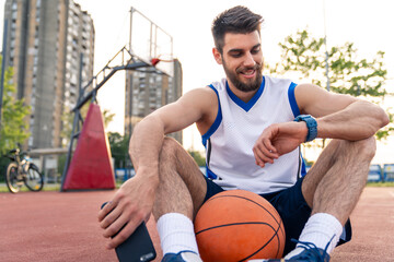 Young Caucasian athlete in white jersey monitors smartwatch during basketball practice on urban outdoor court near residential buildings.