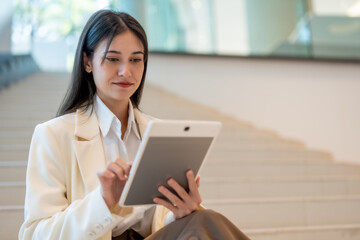 young woman in white blazer sits on stairs, using tablet. She appears focused and engaged, showcasing modern technology in professional setting