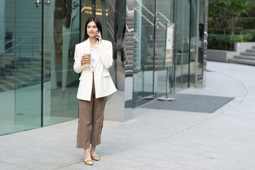 woman in stylish white blazer and brown pants stands outside modern building, holding coffee cup...