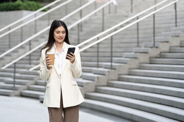woman in white blazer checks her phone while holding coffee cup, standing on outdoor stairs. She...