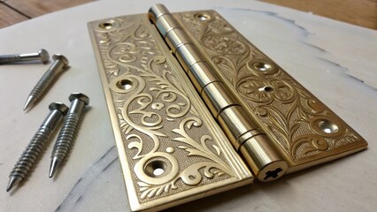 A detailed capture of a shiny decorative hinge featuring unique filigree patterns accompanied by a few wellworn screws resting on the side of a clean textured work surface.