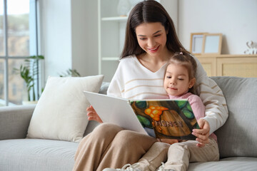 Young mother with her little daughter reading story on sofa at home