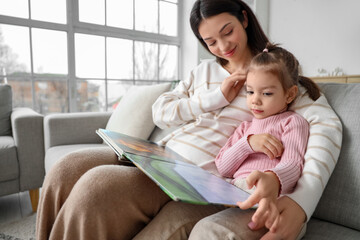 Young mother with her little daughter reading story on sofa at home