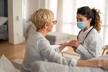 Obraz premium Caucasian female doctor wearing white coat and protective mask provides compassionate medical care to senior patient during professional home healthcare visit.