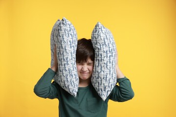 Annoyed boy covering his ears with pillows due to loud sound on yellow background