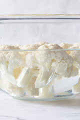 cauliflower florets soaking in a glass bowl of water. On a white marble background