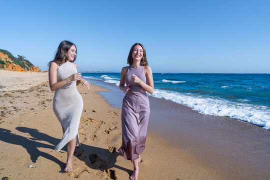 Dos chicas j&oacute;venes elegantes que corren, sonr&iacute;en y se divierten en la playa un sensacional d&iacute;a de verano soleado y con cielo azul.