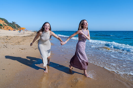 Dos chicas j&oacute;venes elegantes que corren cogidas de la mano, sonr&iacute;en y se divierten en la playa un sensacional d&iacute;a de verano soleado y con cielo azul.