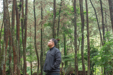 Fototapeta premium A young Asian man traveling alone in the wilderness, hiking through a lush green forest. Capturing the essence of exploration and adventure, perfect for promoting travel and outdoor activities.
