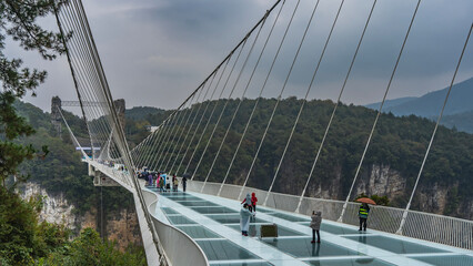 The unique longest suspended glass bridge over the gorge. People in raincoats and carrying...