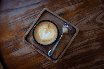 A cup of coffee latte with a heart pattern on foam on a wooden table and a teaspoon with cookies. Top view