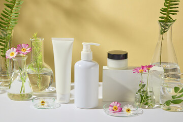 Front view of a set of products features a white tube, pump bottle, and jar mockup, decorated by lab glassware containing tea trees, ferns, green teas, and chamomile branches, in a yellow spa backdrop