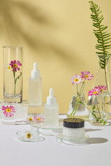 Front view of two dropper bottles and a jar placed on a center on a glass podium, next to some laboratory glassware containing chamomile branches and fern leaves. Set up against a yellow background.