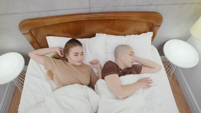 A lesbian woman wakes up in the morning, stretching and yawning, gently waking her bald girlfriend beside her with a loving gesture, starting the day together