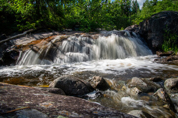 Norwegian mountain river with fast flowing clear and sparkling water and a waterfall that forms white foam on rocks surrounded by green coniferous trees. Sunny summer day.