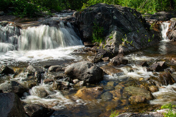 Fototapeta premium Norwegian mountain river with fast flowing clear and sparkling water and a waterfall that forms white foam on rocks surrounded by green coniferous trees. Sunny summer day.
