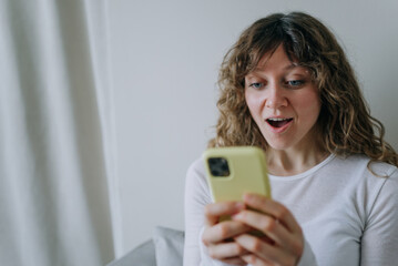 Fototapeta premium Young woman with curly hair holding a yellow smartphone and looking surprised by the news she is reading while sitting on a sofa at home