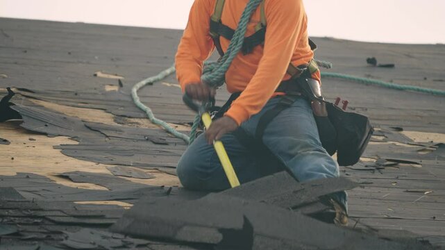 Roofer using tools to strip worn-out shingles while secured with a safety harness.