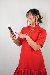 Fototapeta premium Young asian woman wearing red traditional chinese dress is using smartphone with smiling expression, isolated over white background. Concept for Lunar New Year Party.