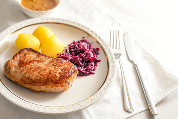Pork loin steak with red cabbage and potatoes on a white plate with cutlery on a light napkin, traditional German meat dish, copy space, selected focus