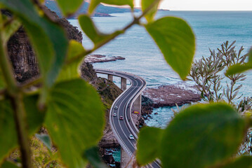 A picturesque view of a coastal bridge, framed naturally by vibrant green leaves. The winding road hugs the rugged cliffs and overlooks the sparkling blue ocean below