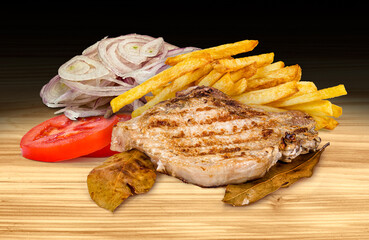 Grilled pork chop with French fries, sliced onion, and tomato on a wooden background. This is a high-resolution, dark scene.