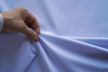 A hand pulling, holding blue delicate fabric with soft folds on light textile background, close up, copy space.
