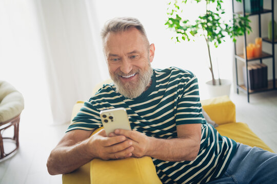 Elderly man joyfully using smartphone while relaxing at home in a sunlit living room - Powered by Adobe