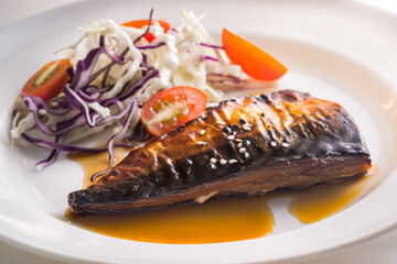 Close-up view of a white plate holds a piece of teriyaki-style grilled mackerel with a light orange-brown teriyaki sauce