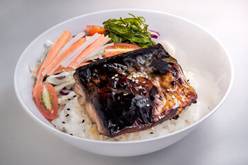 Close-up of a white, round bowl holds a portion of rice topped with a piece of teriyaki-style grilled fish
